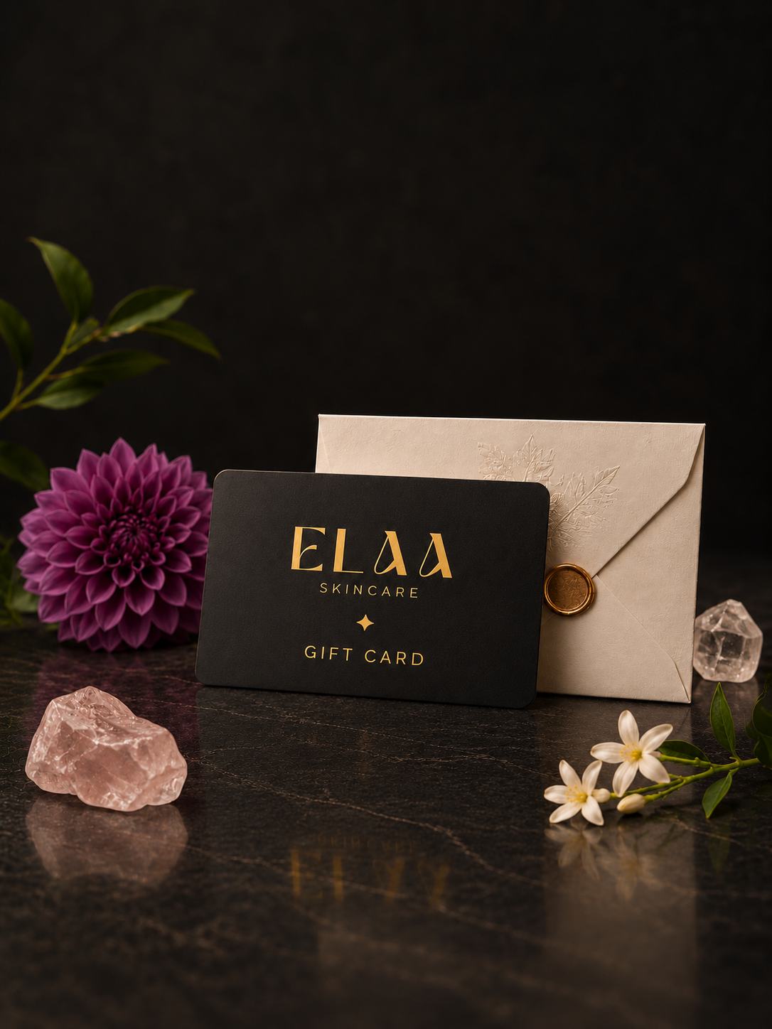 ELAA black and gold gift card with envelope, dahlia, crystals and white flowers on a flat dark surface